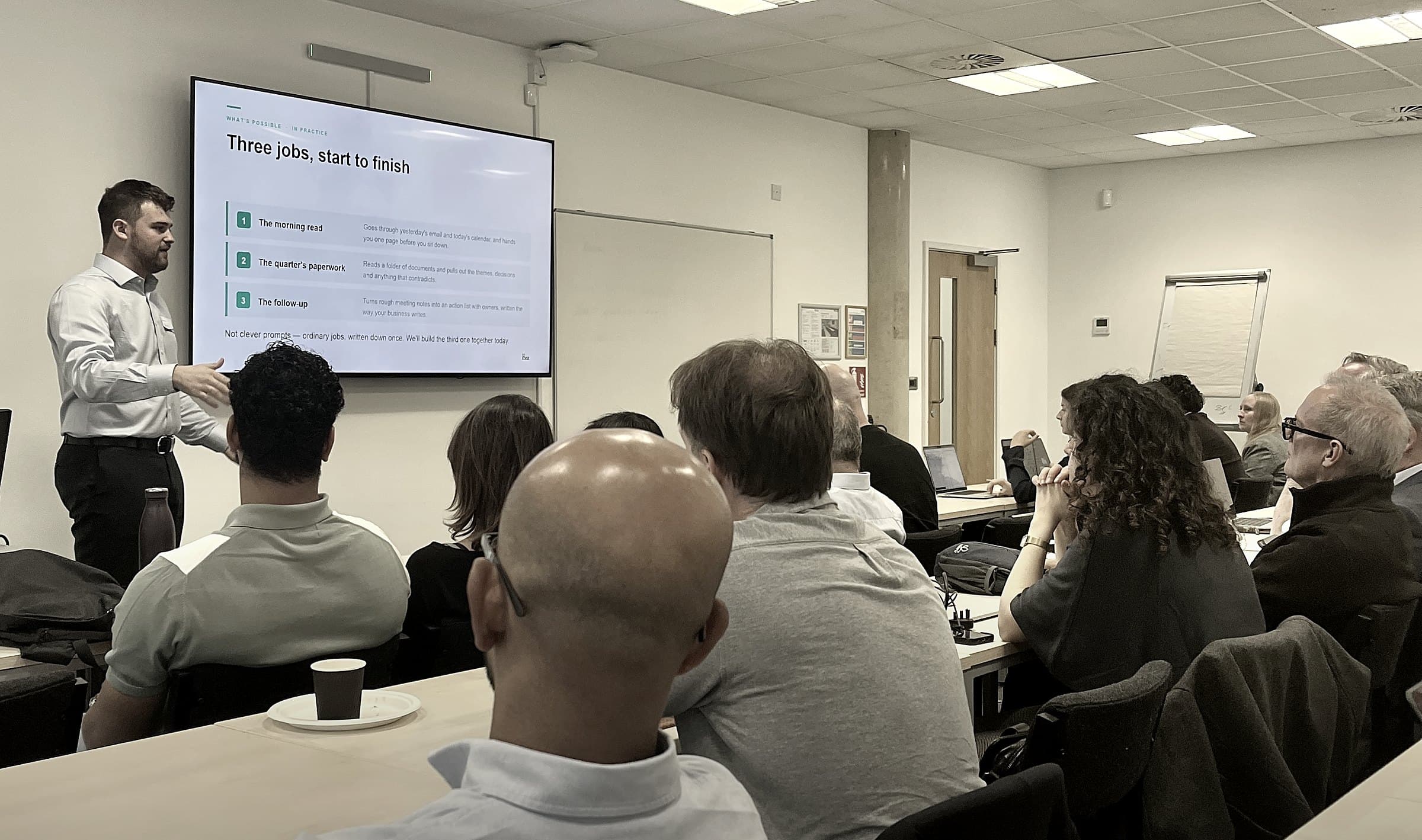 A full room of business owners at an AI Edge session at the Thames Valley AI Hub in Reading, with a slide on screen showing three everyday jobs being worked start to finish